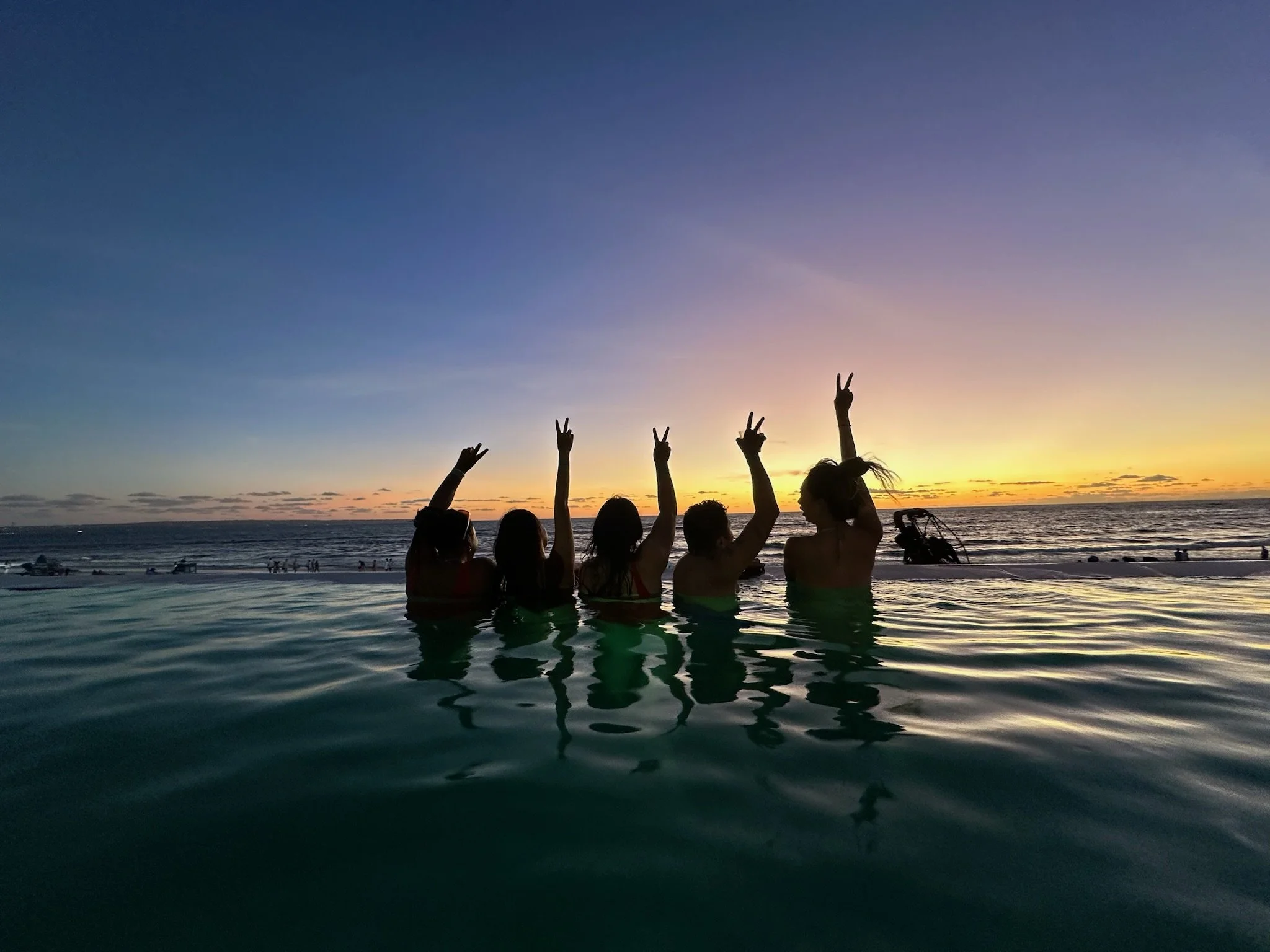 Five people submerged in a pool at sunset making peace signs with their hands, overlooking the ocean.