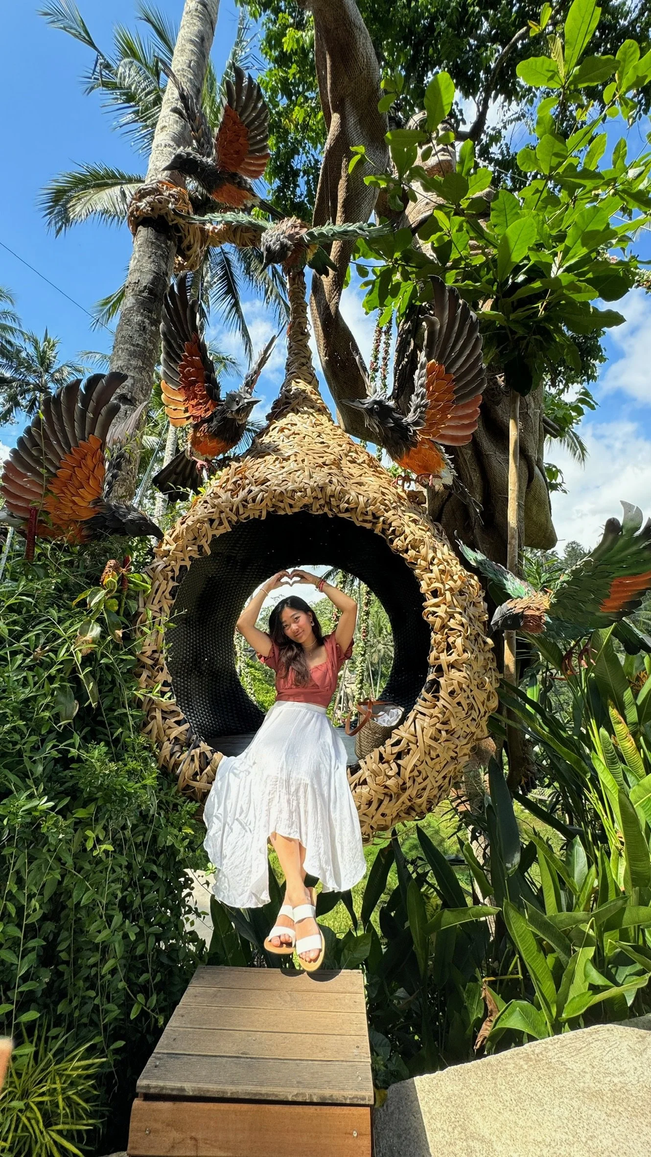 A woman sitting inside a large, circular woven hanging chair, surrounded by lush greenery with palm and broad-leaf trees, under a blue sky with some clouds. The woman is wearing a pink top, white skirt, white sandals, and is posing with her arms abov