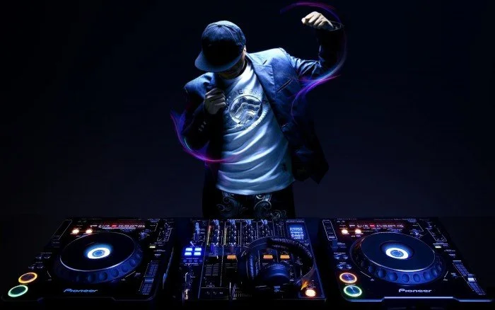 Person DJing with mixing equipment in a dark setting, wearing a cap, jacket, and t-shirt, with colorful light effects.
