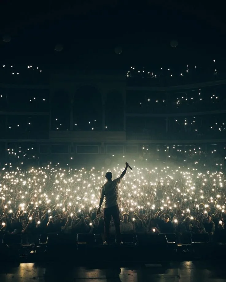 Performer on stage holding a microphone towards a large audience in a concert hall, with many audience members holding up their phones with flashlights on.
