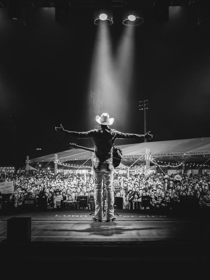 A cowboy on stage with arms outstretched, facing a large crowd at a concert or event, under bright stage lights in a black-and-white photo.