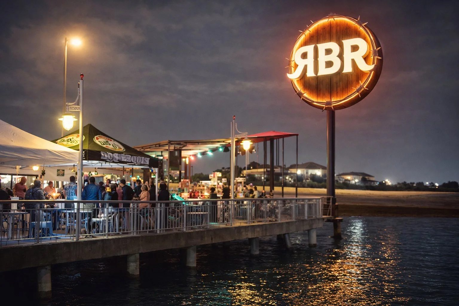 Nighttime scene at a waterfront restaurant with people dining under tents, illuminated by string lights, and a large circular sign with the letters 'YBR' glowing brightly.