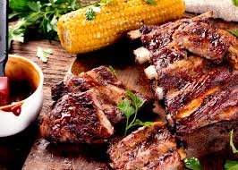 Grilled steaks alongside corn on the cob, with a cup of barbecue sauce on a wooden table.