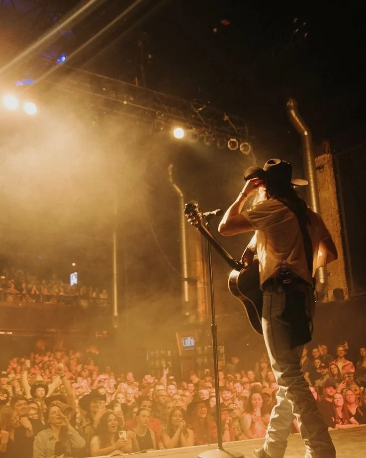 Performer with a guitar on stage in front of a large audience, illuminated by stage lights in a concert venue.