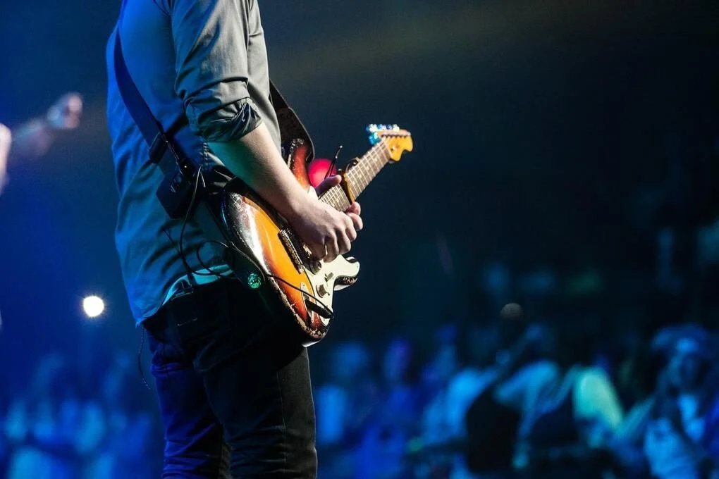 Person playing electric guitar on stage during a concert with a crowd in the background.