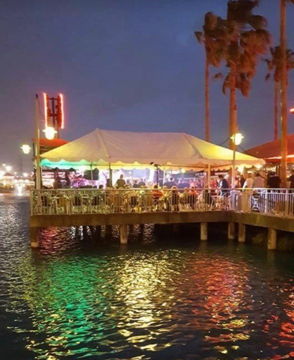 A lively outdoor restaurant at night on a pier over water, with a large tent, colorful lights, and palm trees in the background.