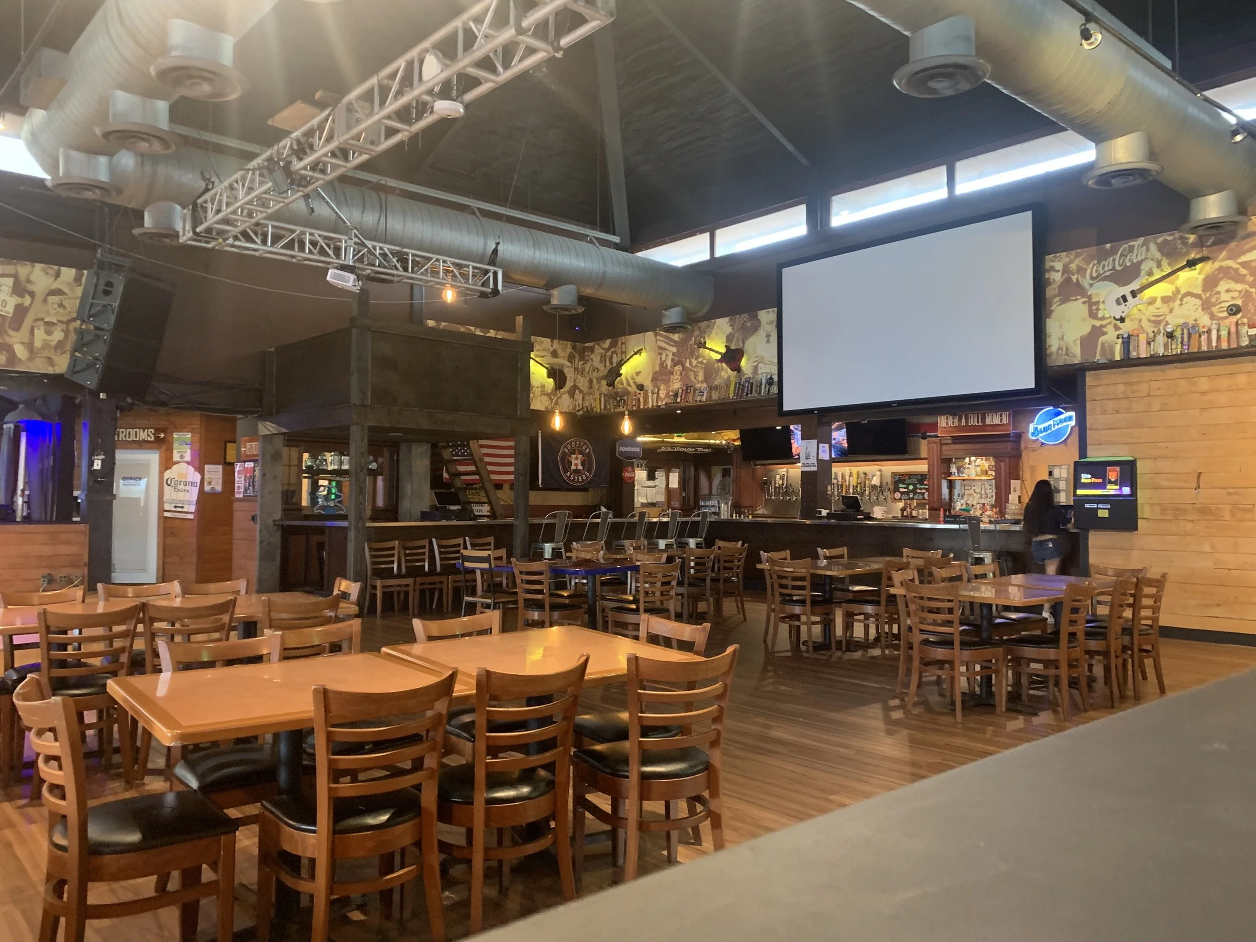 Interior of a bar or restaurant with wooden tables and chairs, a bar counter with drinks, large screen TVs, and industrial decor including exposed ductwork and wall art.