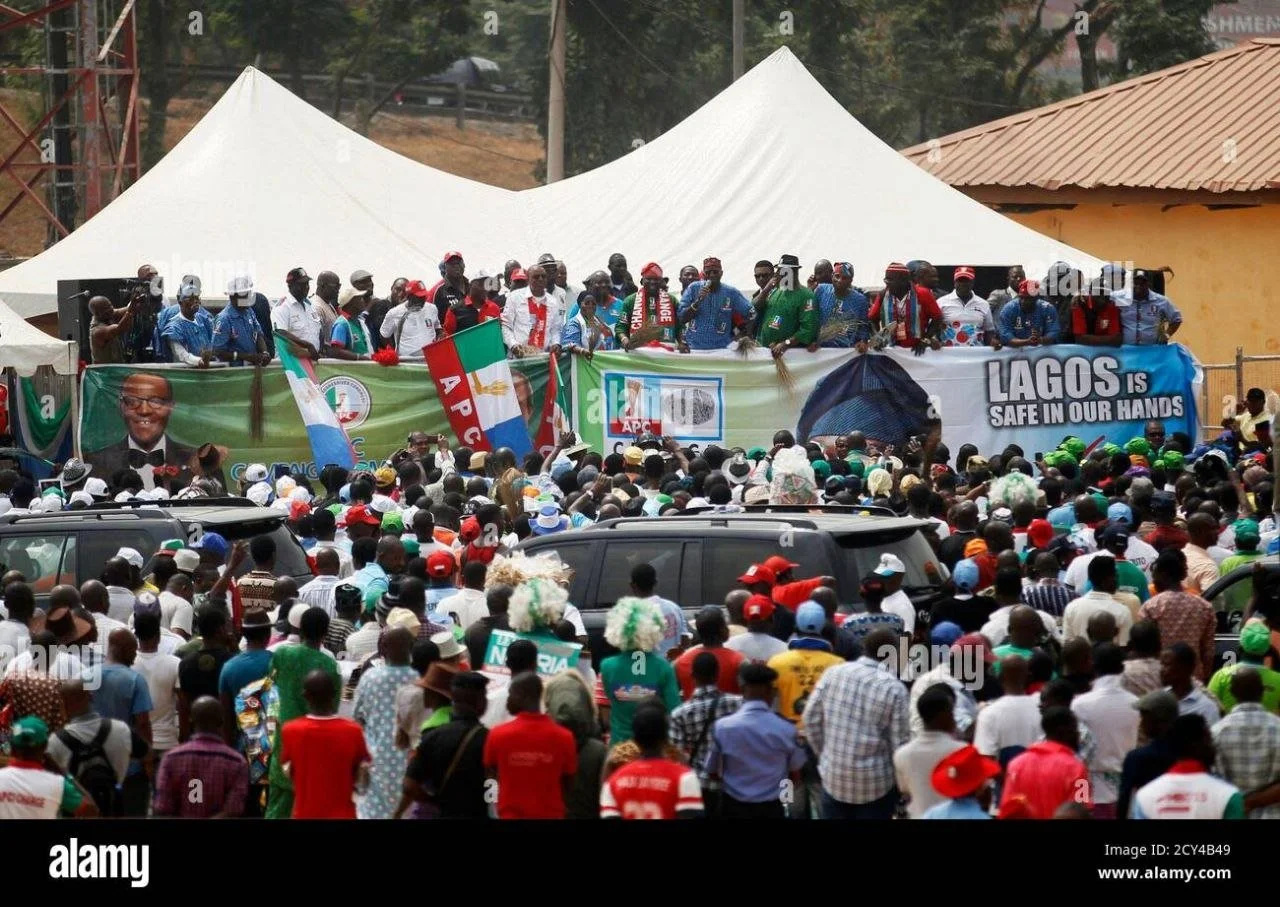 A large crowd gathered at a political rally with many people wearing hats and shirts in black, white, red, and green. Politicians are on a stage behind a banner that reads 'LAGOS IS SAFE IN OUR HANDS,' with flags and a large photograph of a man. The 