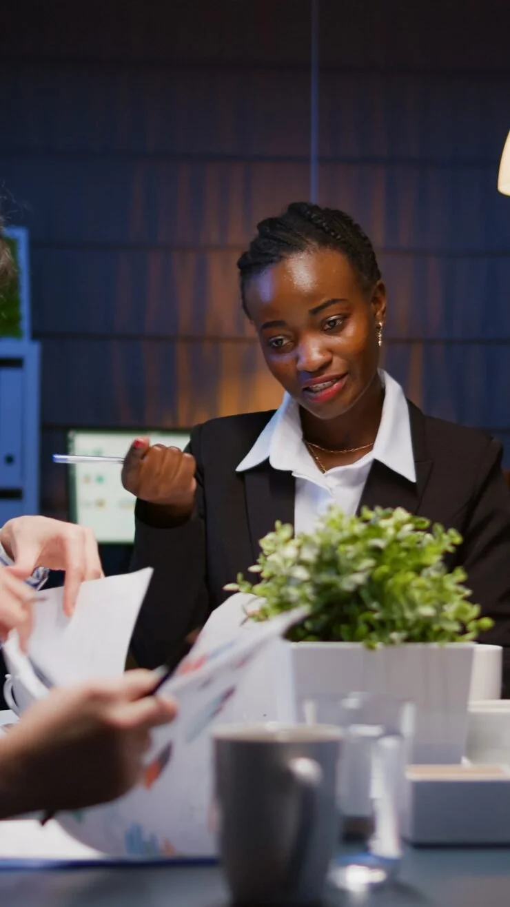 A woman in a white shirt and black blazer sitting at a table with a potted plant, talking to someone and holding a pen, in a modern office or conference room setting.