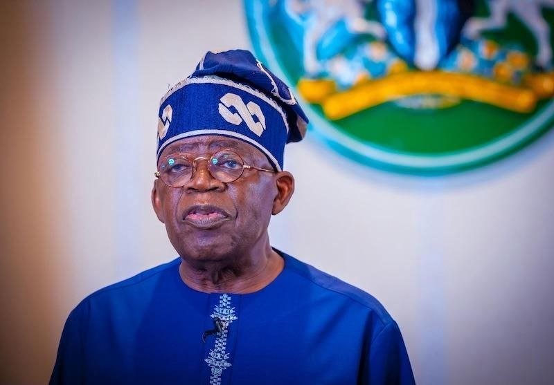 An elderly man wearing glasses, a blue traditional outfit, and a matching traditional hat, standing indoors with a blurred emblem or flag in the background.