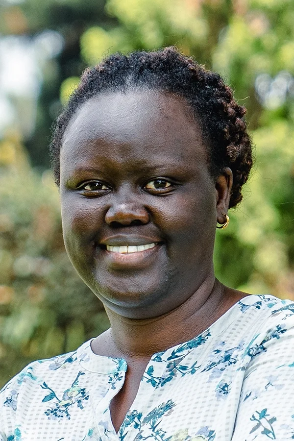 A woman with dark skin and short, curly black hair smiling outdoors with trees in the background.