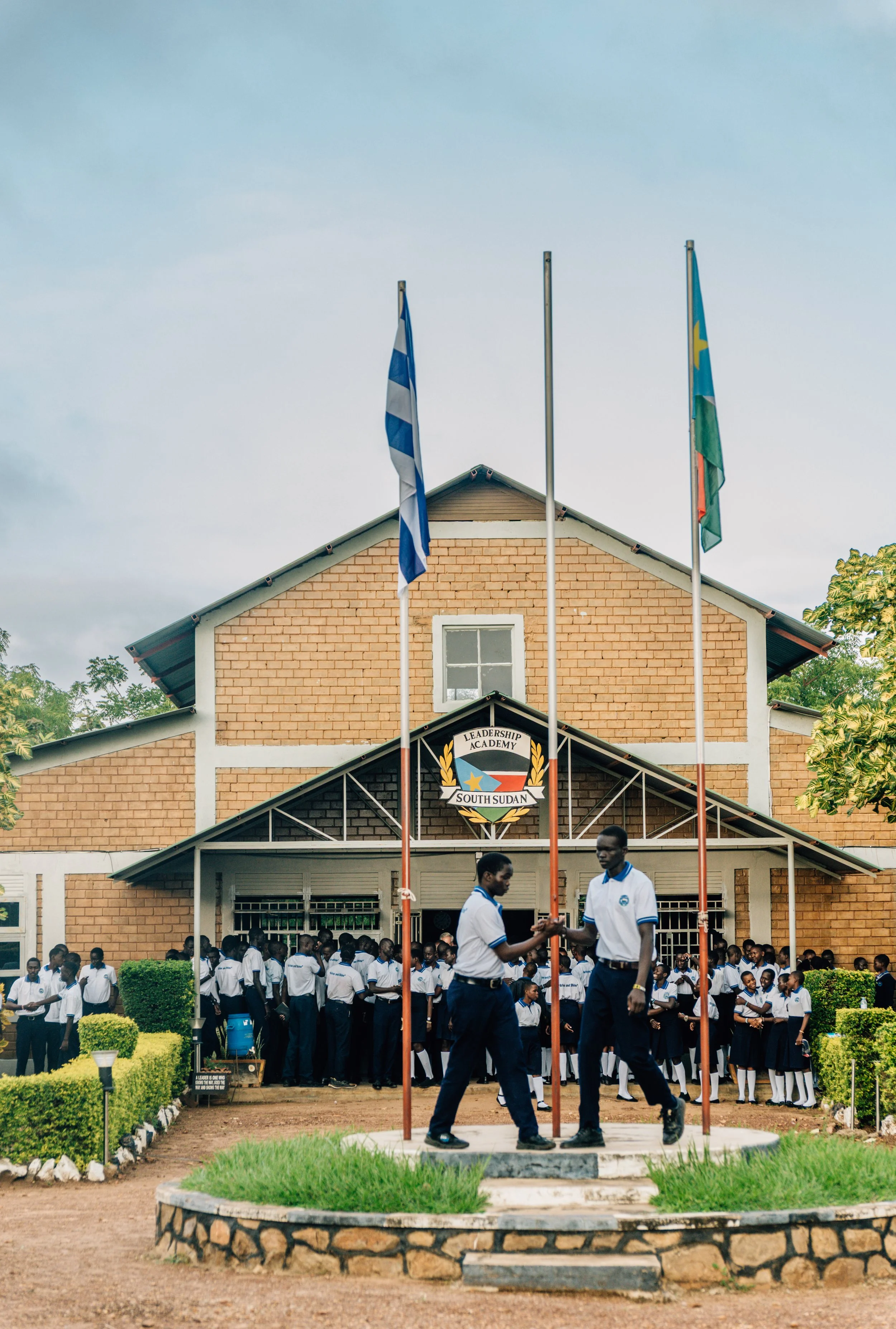School students gathered outside a brick building at Leadership Academy in South Sudan, with two students raising a flag as others watch.