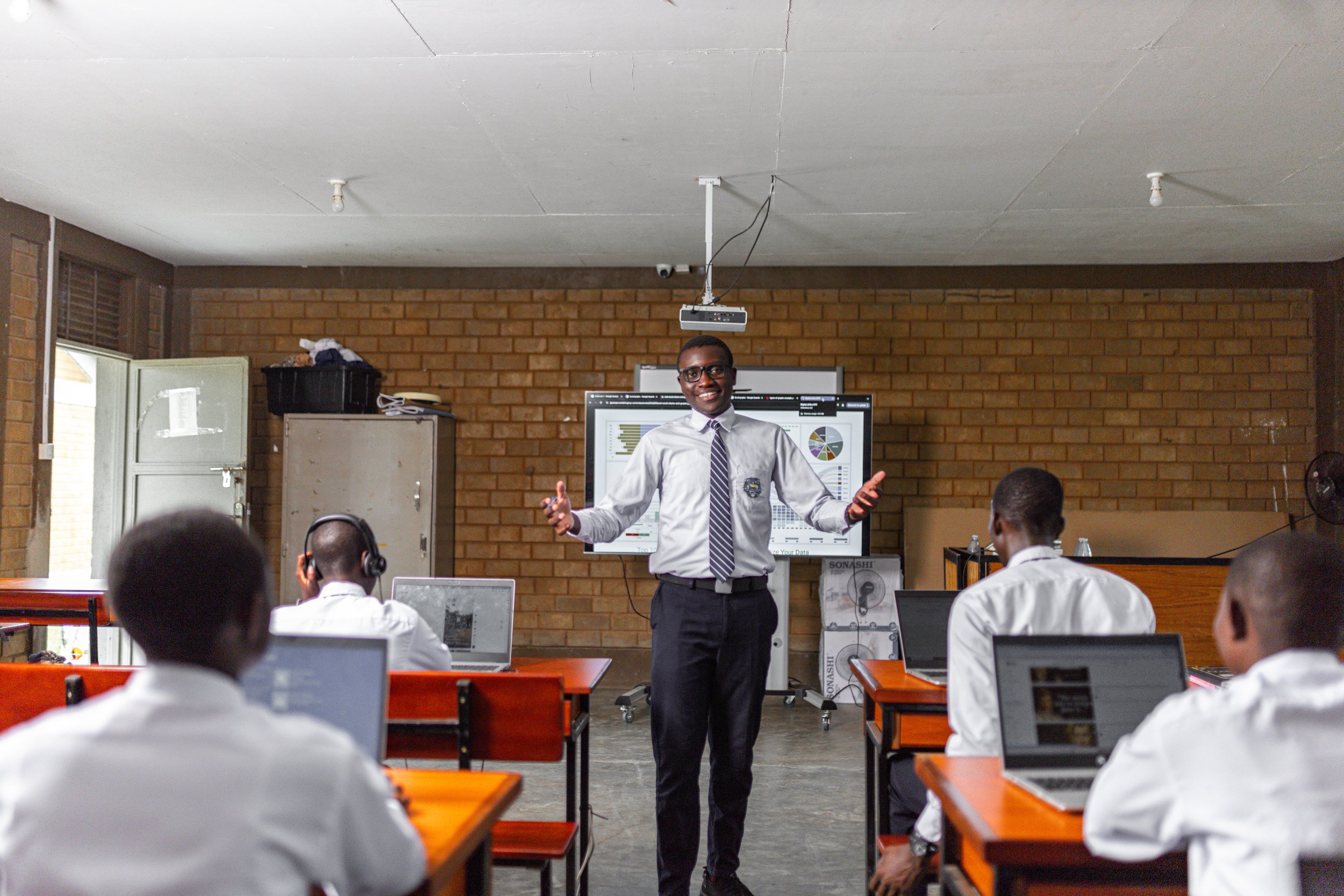 A classroom with students working on laptops and a teacher standing in front of a digital screen giving a lesson.