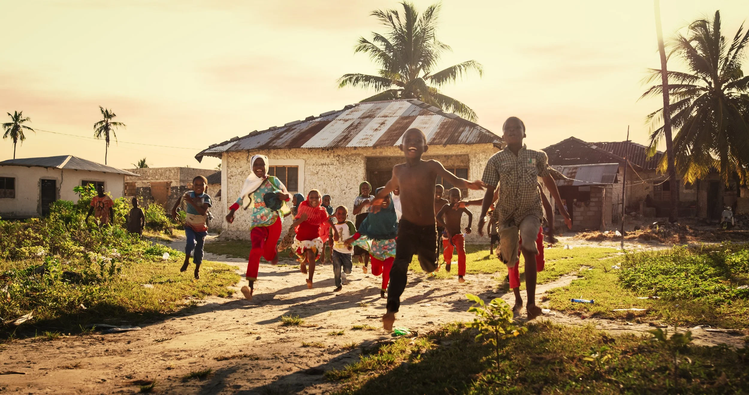 Children running and playing outdoors in a village with rustic buildings and palm trees at sunset.