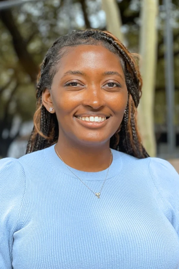 A woman with dark hair styled in braids, smiling and wearing a light blue top and a necklace, outdoors with trees in the background.