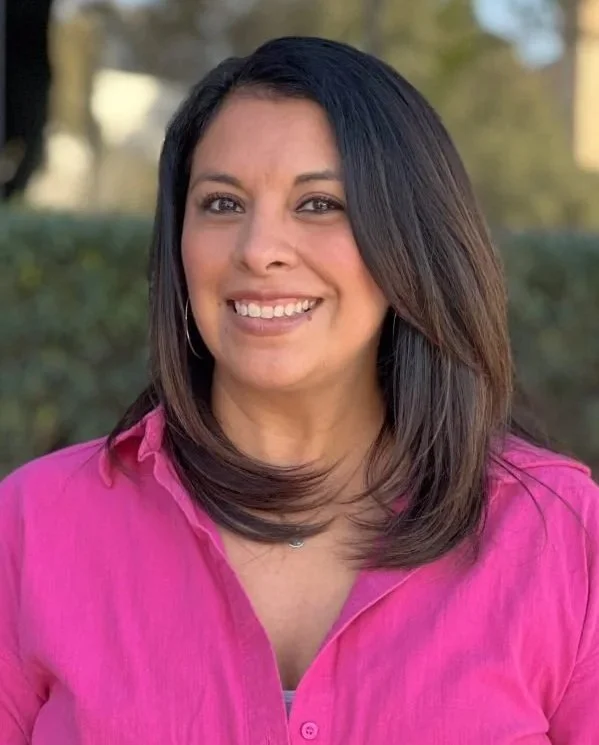 A woman with shoulder-length dark hair, smiling, wearing a pink button-up shirt outdoors.
