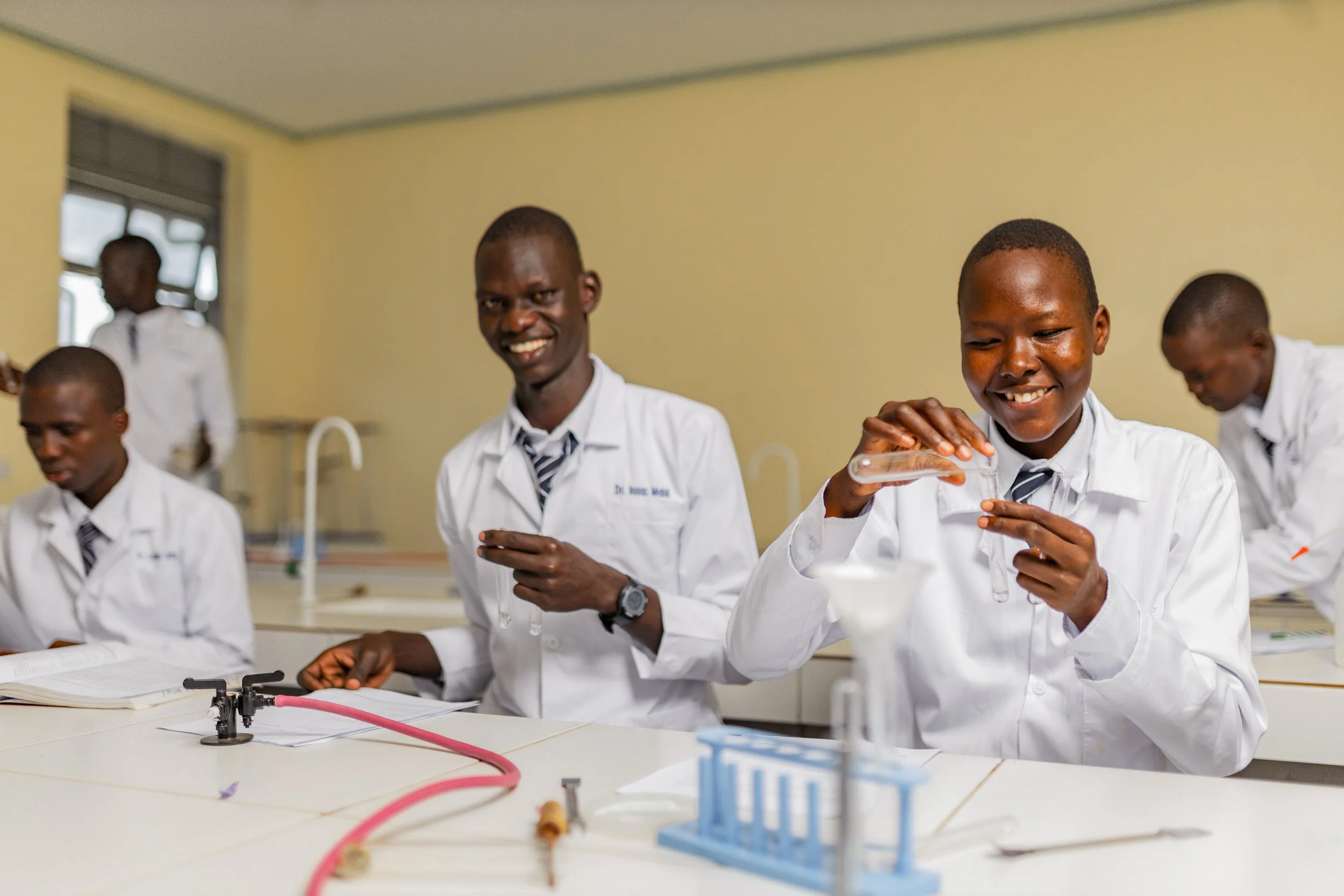 Students in a science classroom conducting experiments with laboratory equipment, smiling and wearing white lab coats.