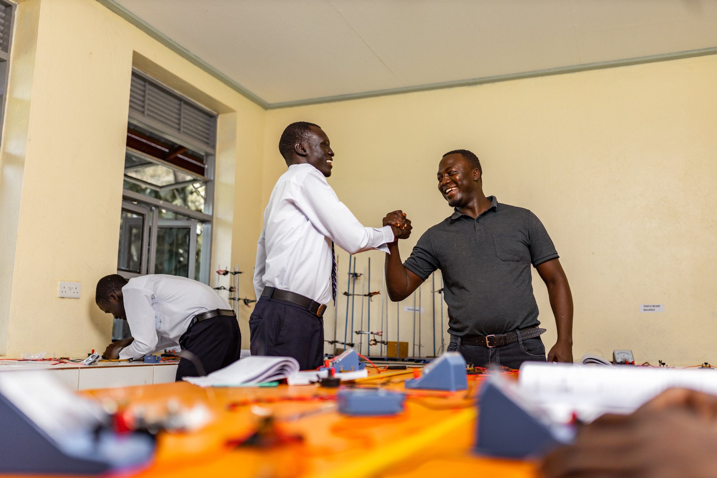 Two men smiling and shaking hands in a classroom or workshop setting, with a third man working at a table in the background. The scene suggests a successful collaboration or celebration.