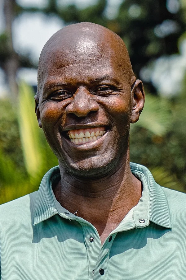 A smiling African American man with a bald head, wearing a light green polo shirt, standing outdoors with blurred green foliage in the background.