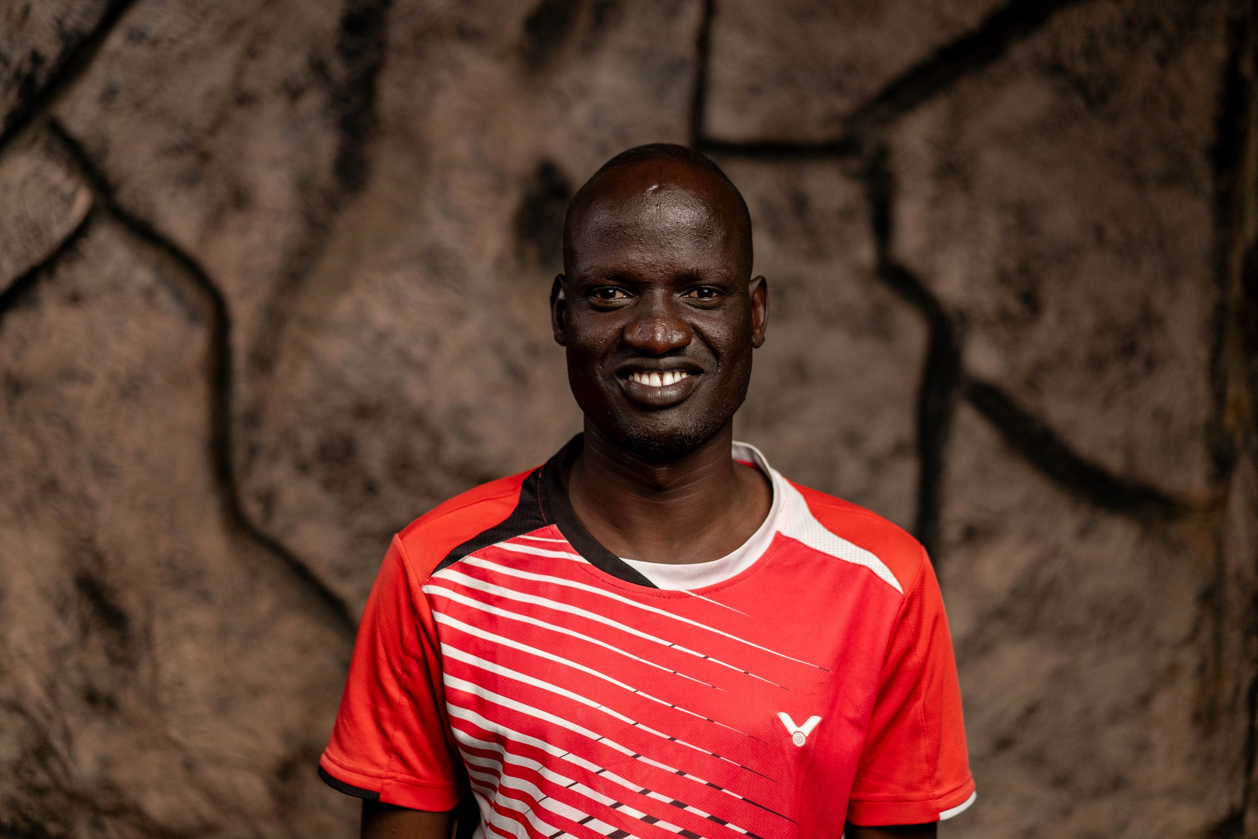 A man in a blue polo shirt with a logo reading 'AYLF Nkumba Chapter' stands in front of a stone wall.
