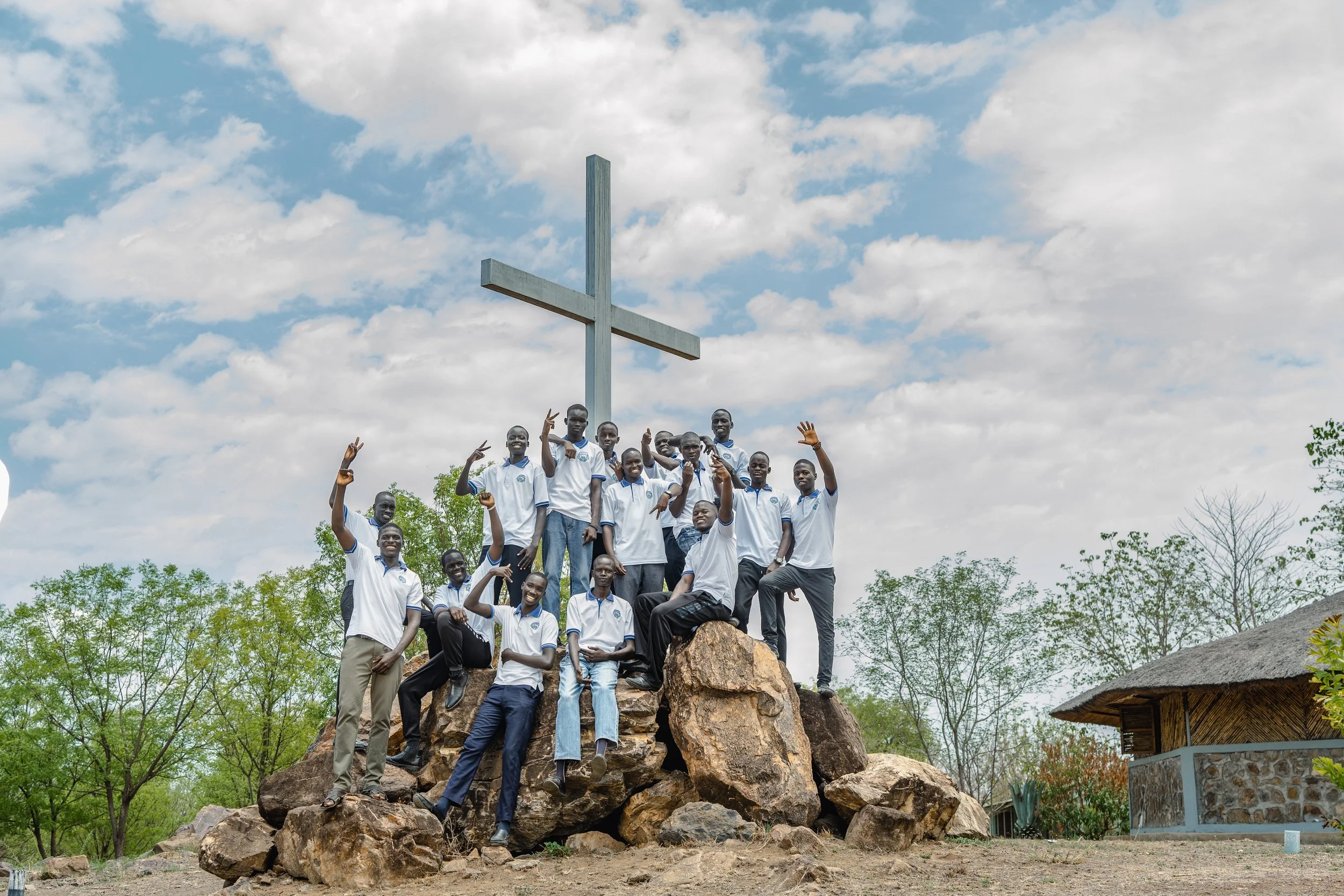 A group of young men in white shirts and dark pants posing on large rocks around a tall cross in an outdoor setting with trees and a small thatched-roof building visible in the background.