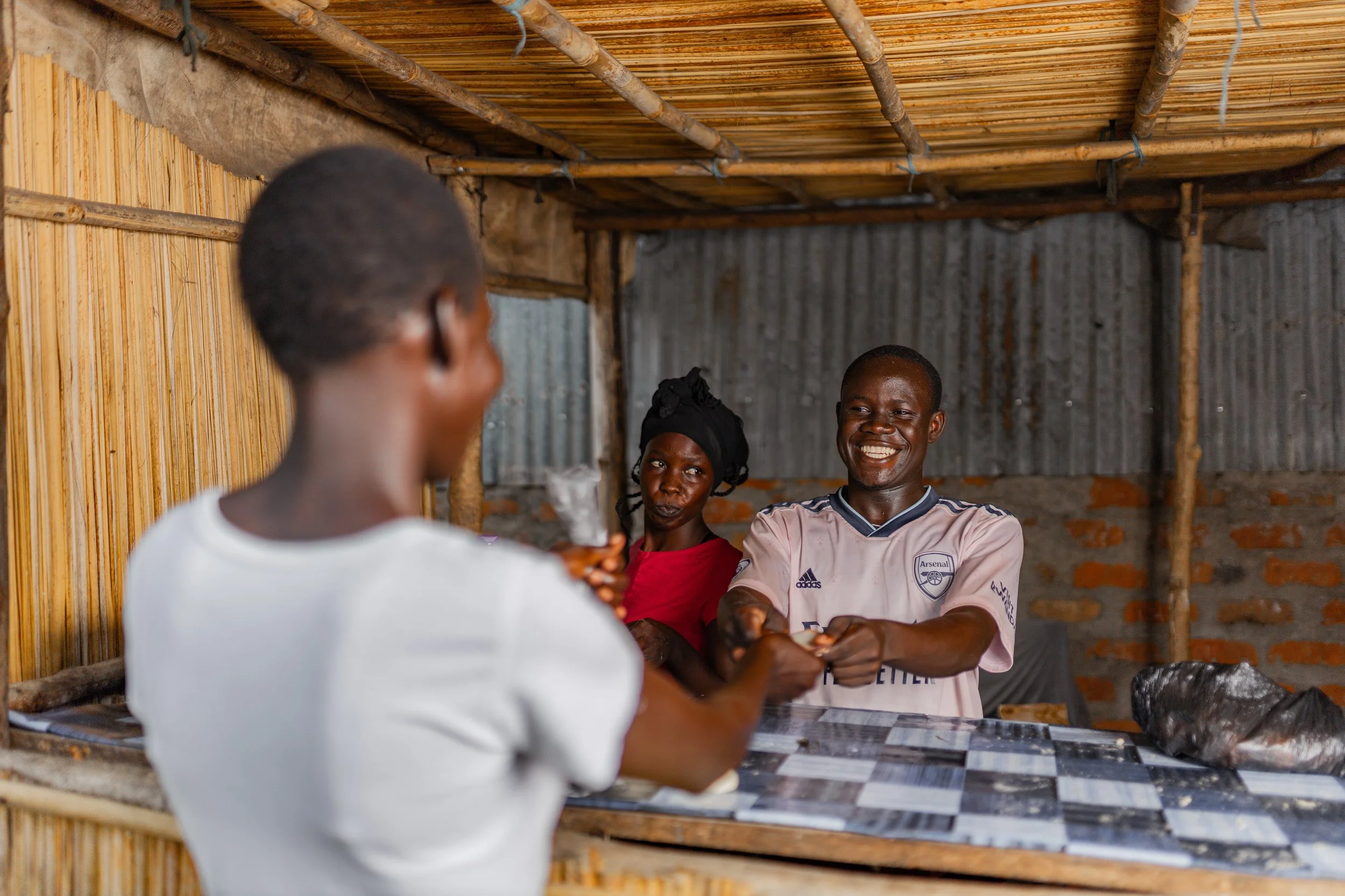 Three people inside a rustic hut with bamboo and corrugated metal walls, smiling as they exchange money.