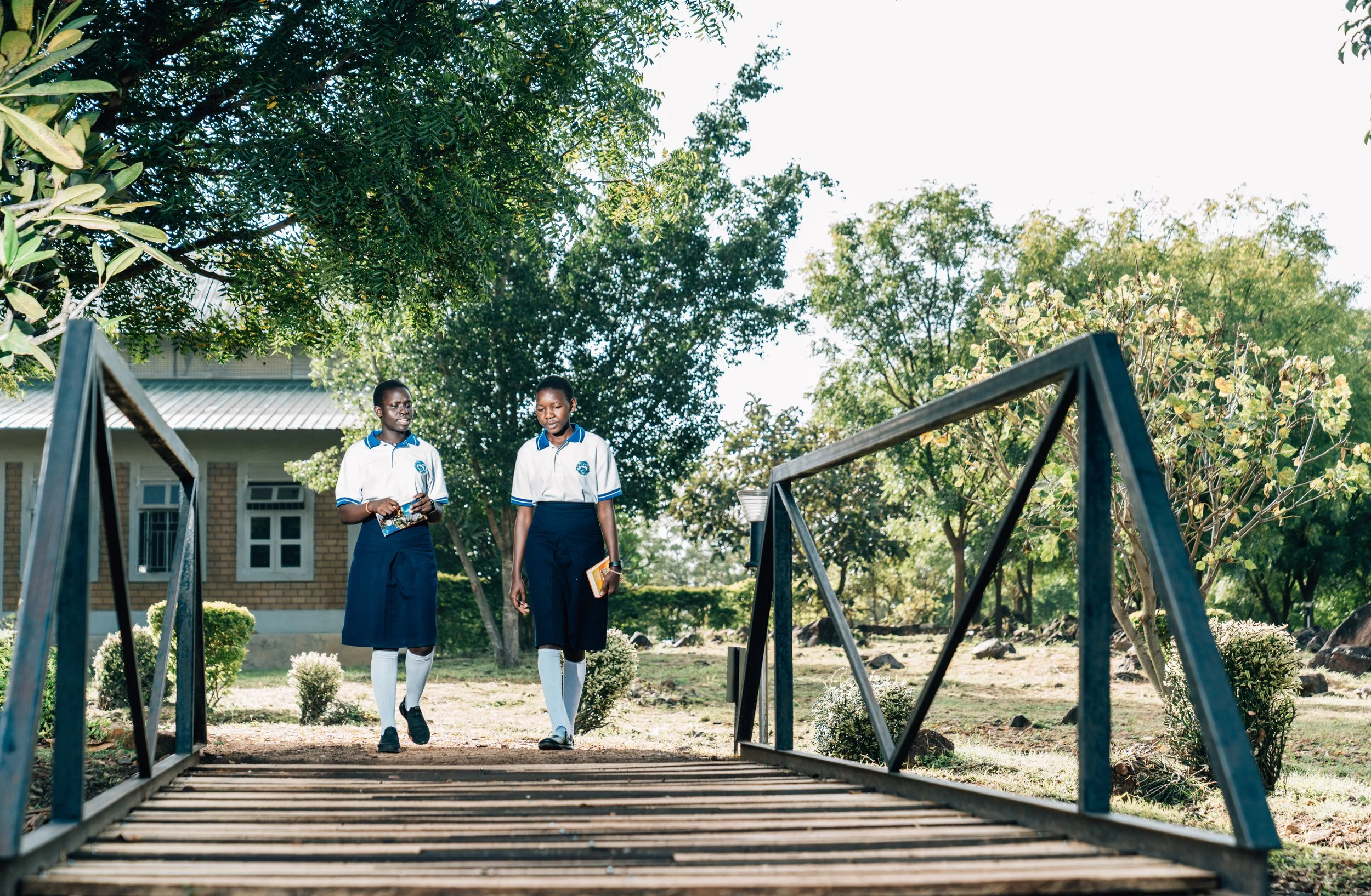 Two students in school uniforms walking on a path in a green outdoor area with trees and a building in the background.