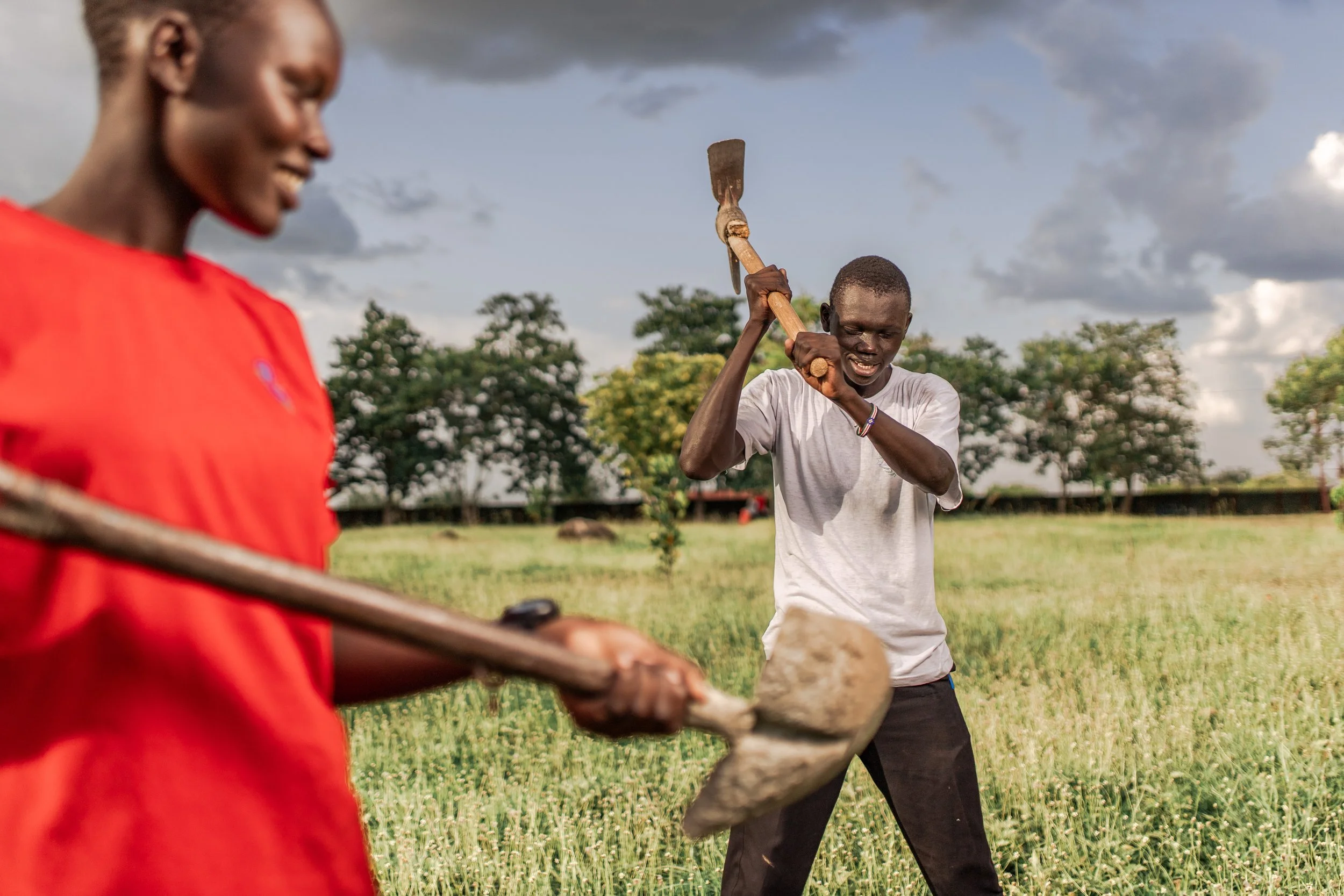 Two young men playing a game of whack-a-mole outdoors in a grassy field with trees and clouds in the background.