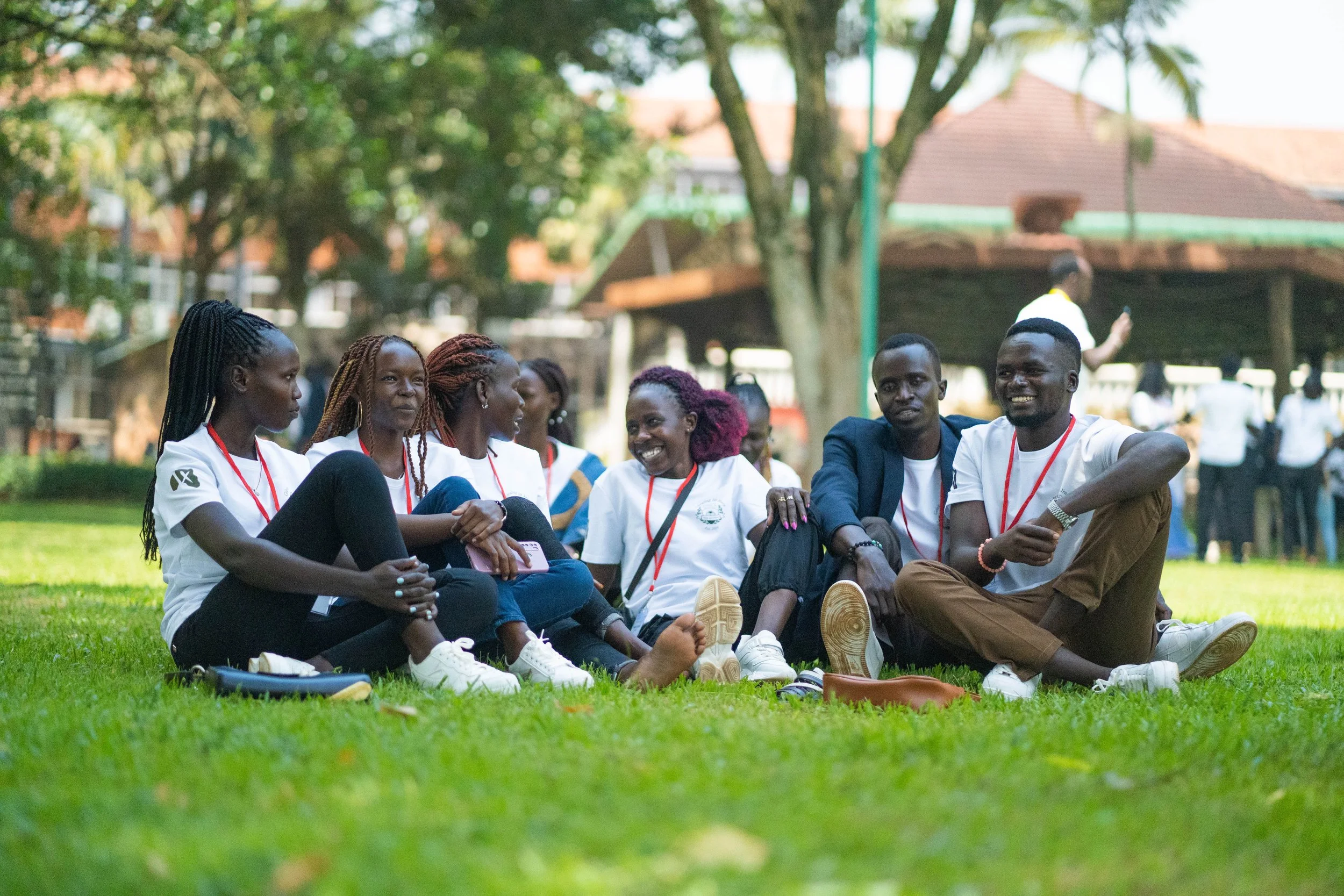 Group of eight young adults sitting on grass outdoors, engaging in conversation and smiling, under trees with a pavilion in the background.