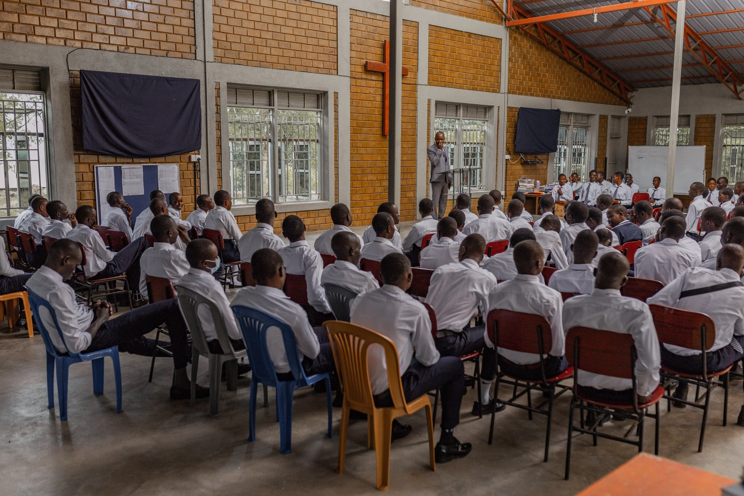A classroom filled with students in white shirts and ties, sitting on chairs, facing a teacher standing at the front near a whiteboard. The room has large windows, brick walls, and a high ceiling with exposed beams.