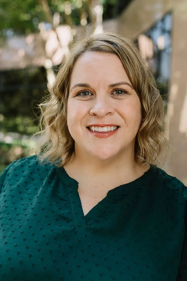 A woman with shoulder-length curly blonde hair smiling outdoors, wearing a dark green textured blouse with a V-neck.