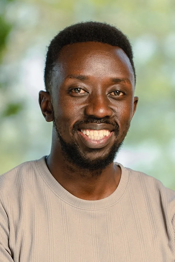 Smiling man with short black hair and a beard, wearing a light gray shirt, in front of a blurred green background.