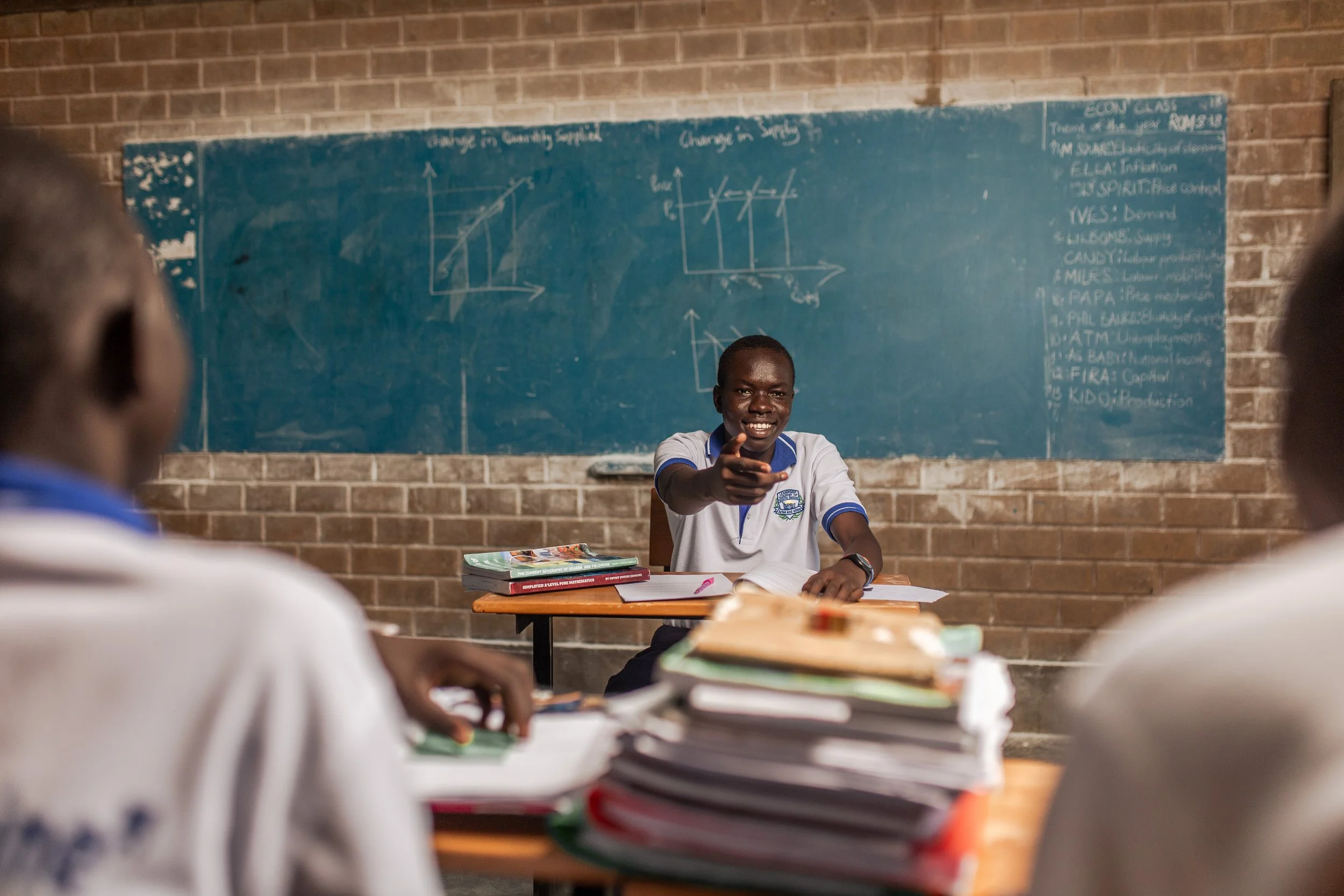 A classroom scene with a smiling teacher pointing at students. The teacher is sitting at a desk with textbooks. Behind him is a chalkboard with diagrams and notes. Students' heads are visible in the foreground.