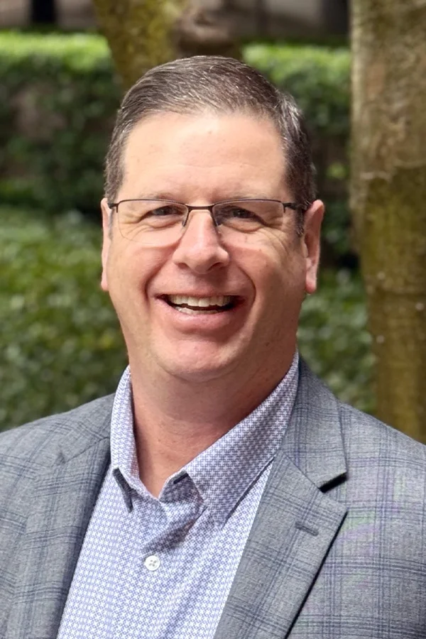 A smiling man with short dark hair, glasses, wearing a gray checked blazer and a collared shirt, standing outdoors with trees and greenery in the background.