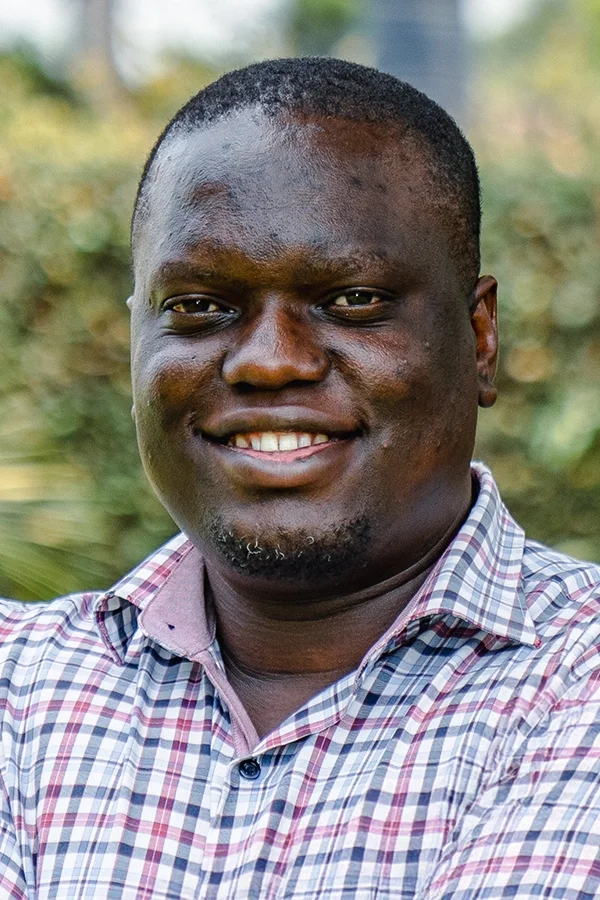 Portrait of a smiling black man with short hair, wearing a checkered shirt, outdoors with blurred greenery in the background.