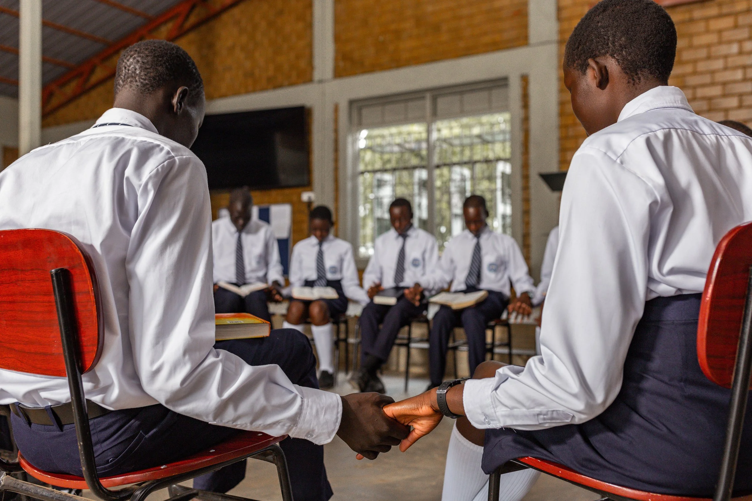Two students holding hands in a school setting with other students sitting in the background.