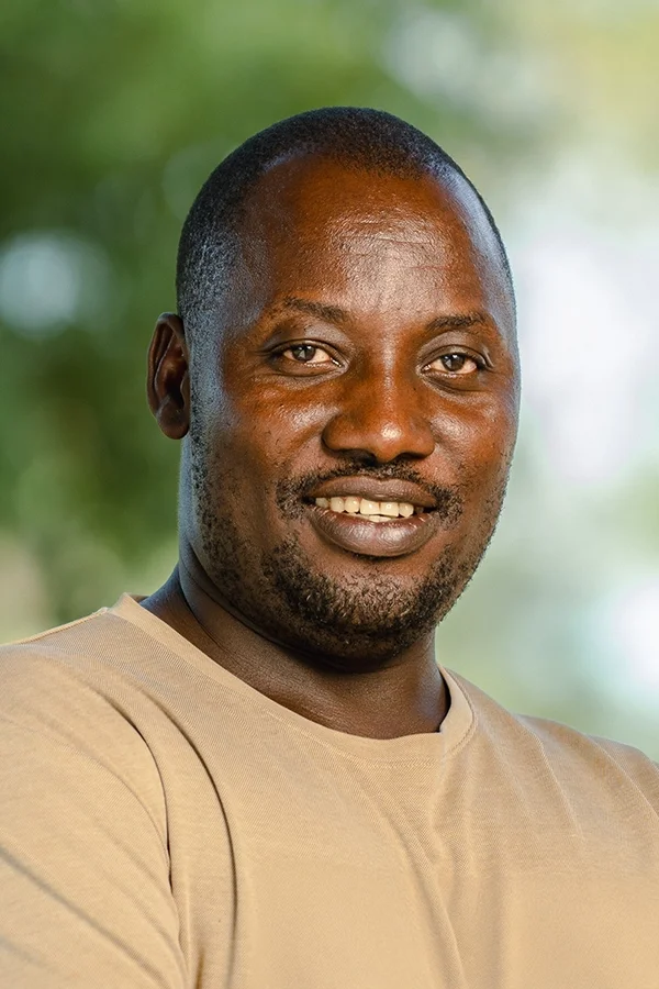 A smiling man with short hair and a beard wearing a beige t-shirt, standing outdoors with blurred green trees in the background.