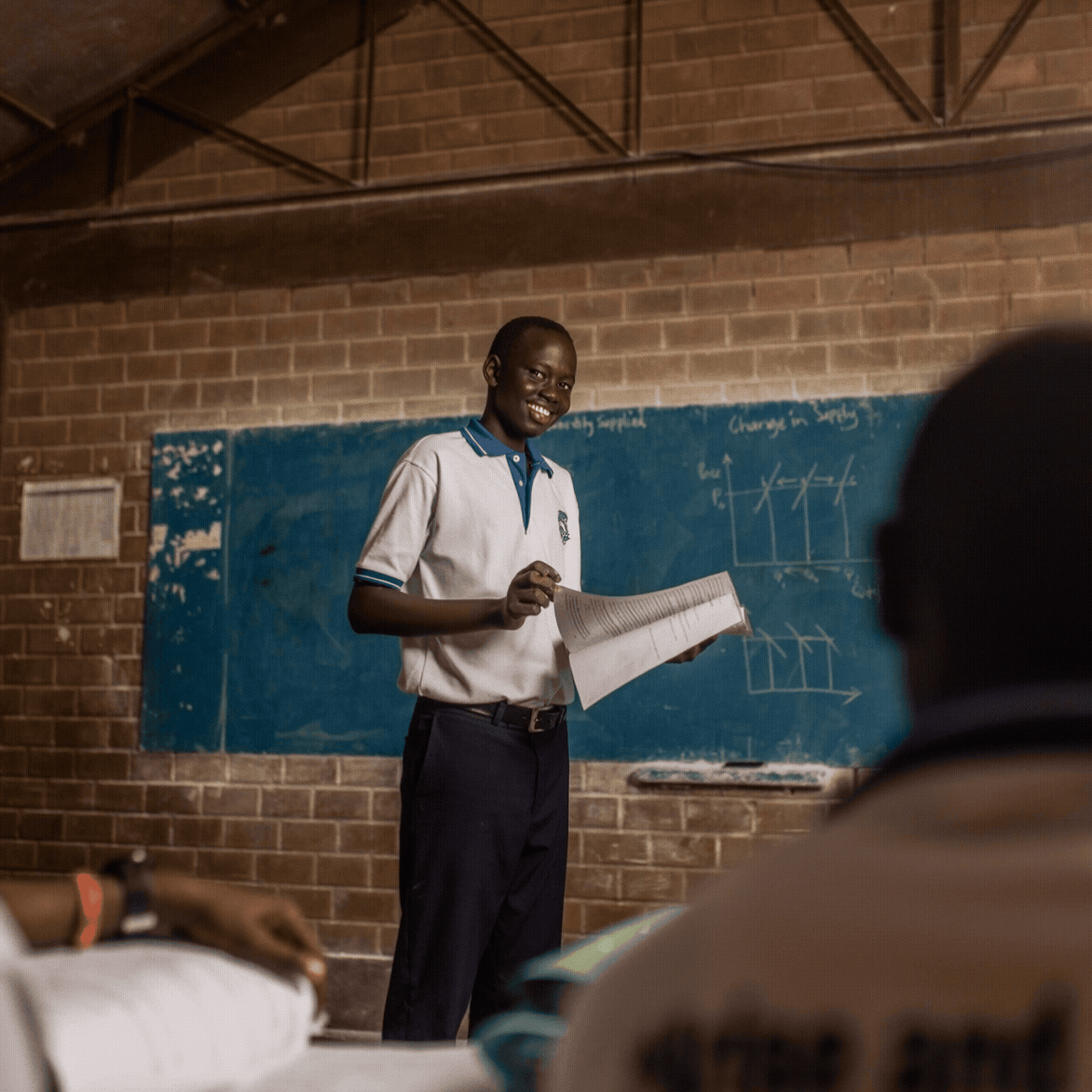 A male teacher smiling and holding a book in a classroom with a chalkboard and diagrams in the background, and students seated in front.