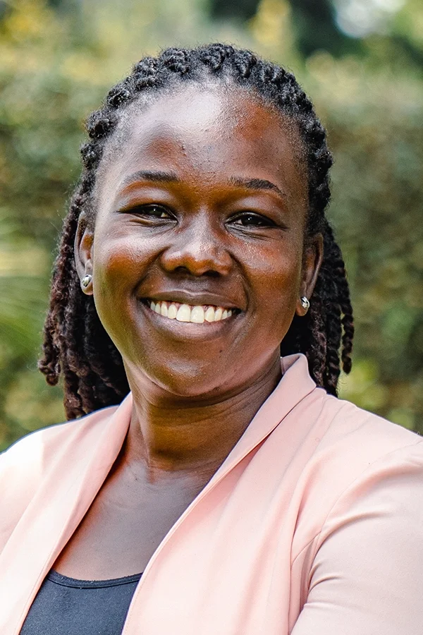 A smiling woman with dark braided hair wearing a light pink blazer and earrings outdoors.