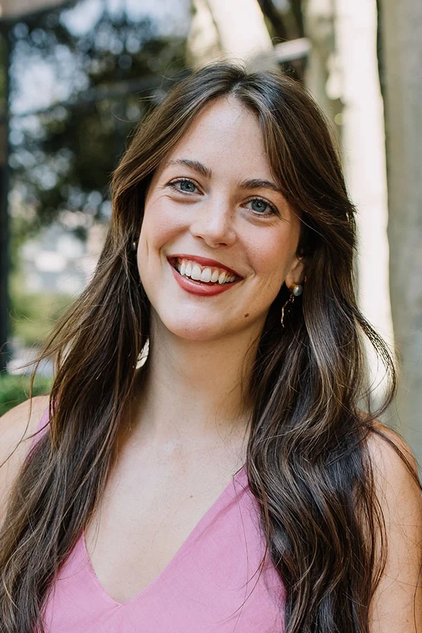 A smiling young woman with long brown hair wearing a pink sleeveless top and pearl earrings outdoors.