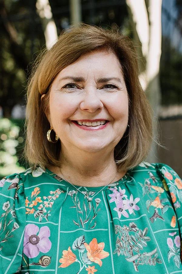 A woman with shoulder-length brown hair smiling, wearing a green floral patterned top, gold hoop earrings, and a silver necklace, outdoors with trees and a modern building in the background.