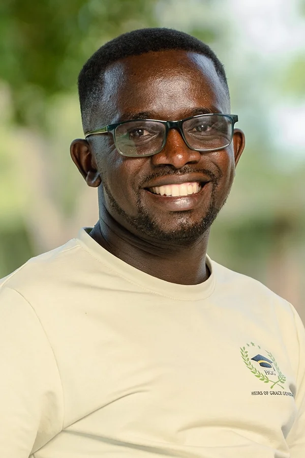 A smiling Black man wearing glasses and a white T-shirt with a logo, outdoors with green trees in the background.