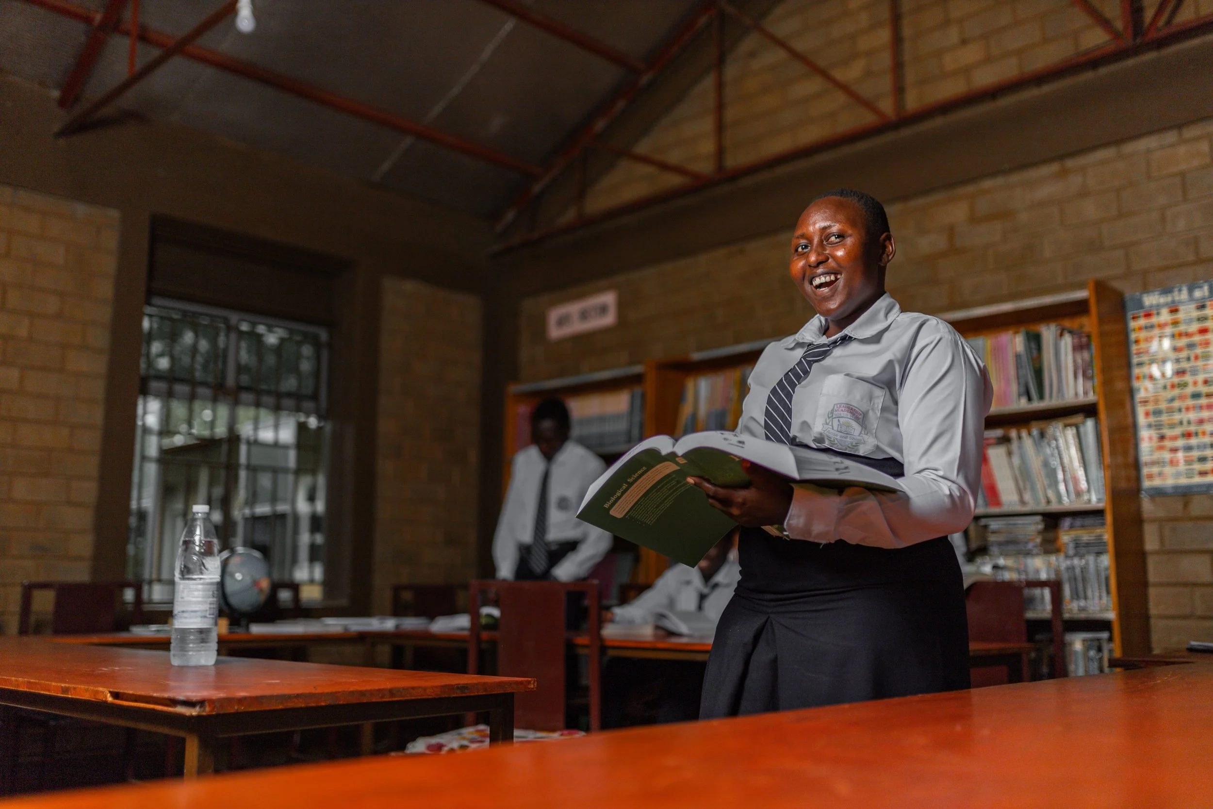 A smiling woman in a school uniform holding an open book in a classroom with a bookshelf, another student, and a bottle of water on the table nearby.
