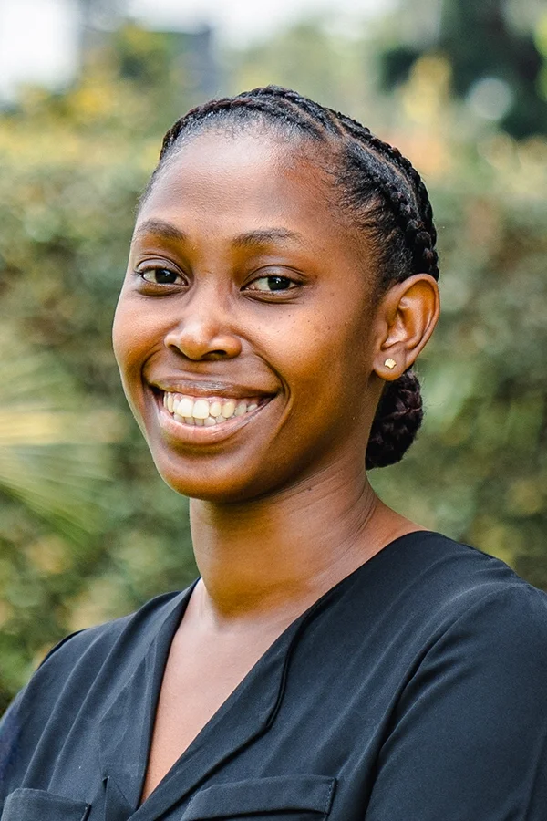 A smiling African American woman with braided hair, wearing a black top, standing outdoors with blurred greenery in the background.