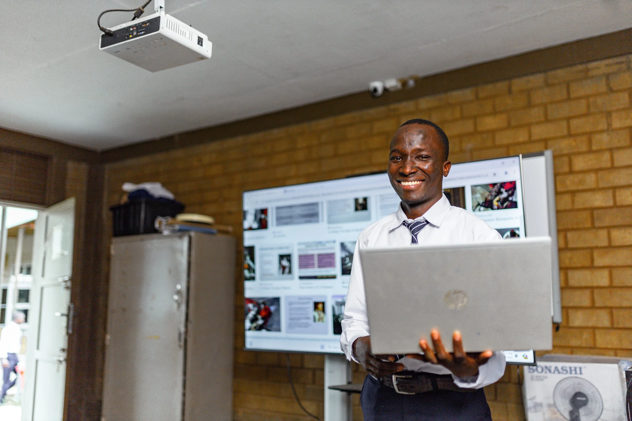 A man in business attire smiling while holding a laptop in a classroom or conference room with a large screen displaying documents behind him.