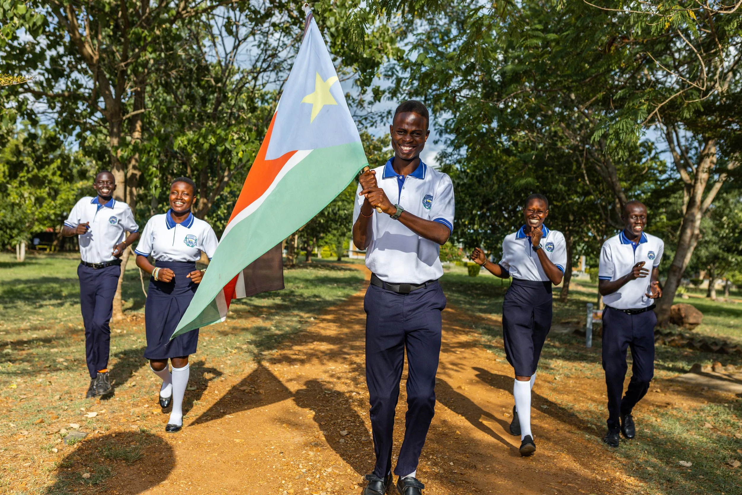 Young students in school uniforms running outdoors while one girl carries the flag of South Sudan, smiling and enjoying a lively activity on a sunny day.
