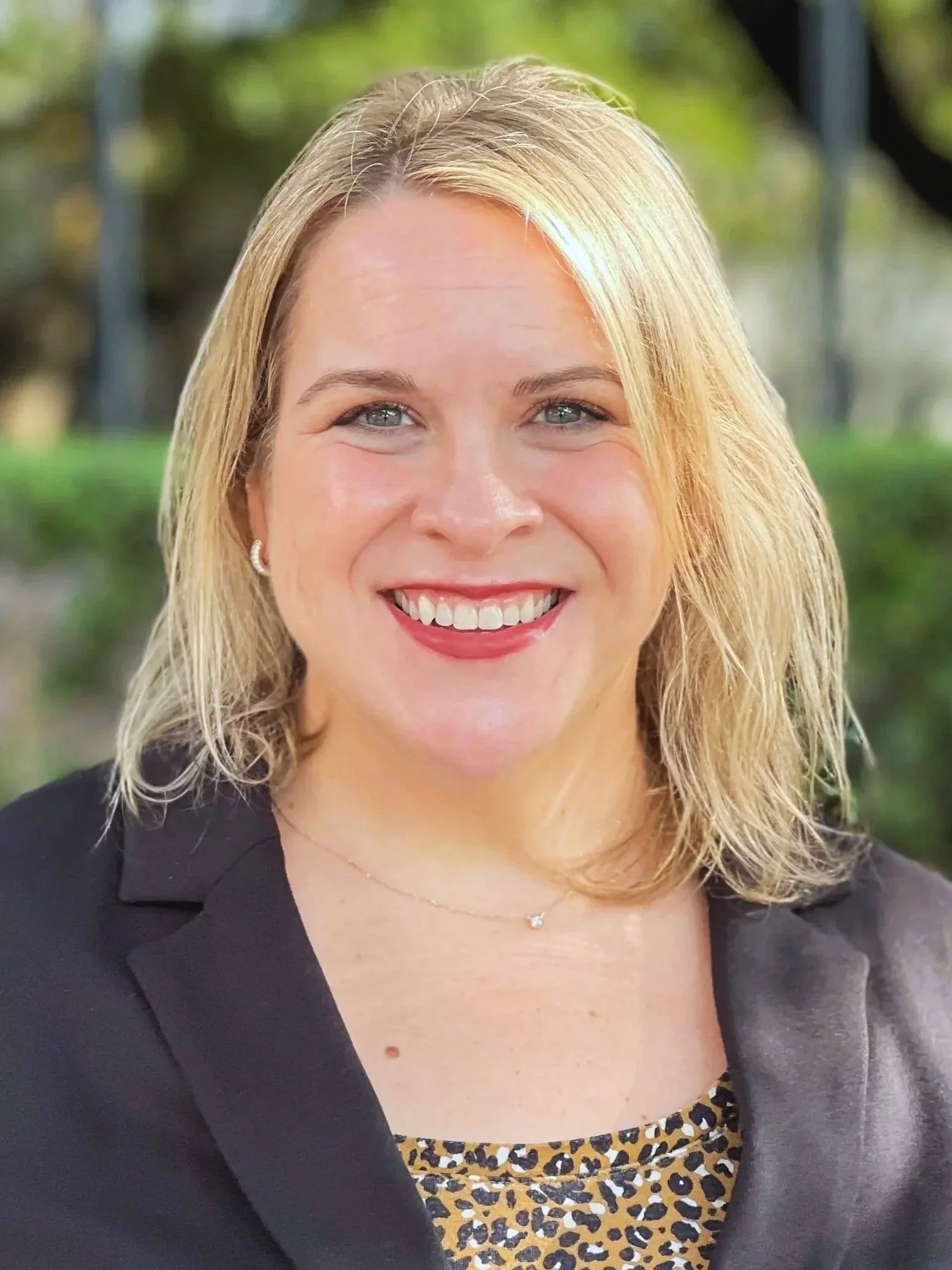 A woman with shoulder-length curly blonde hair smiling outdoors, wearing a dark green textured blouse with a V-neck.