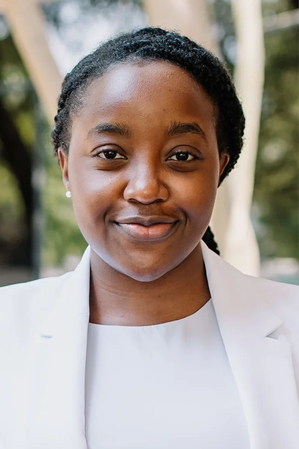 A young woman with dark hair styled in twists, smiling, wearing a white blazer and top, outdoors with blurred greenery in the background.