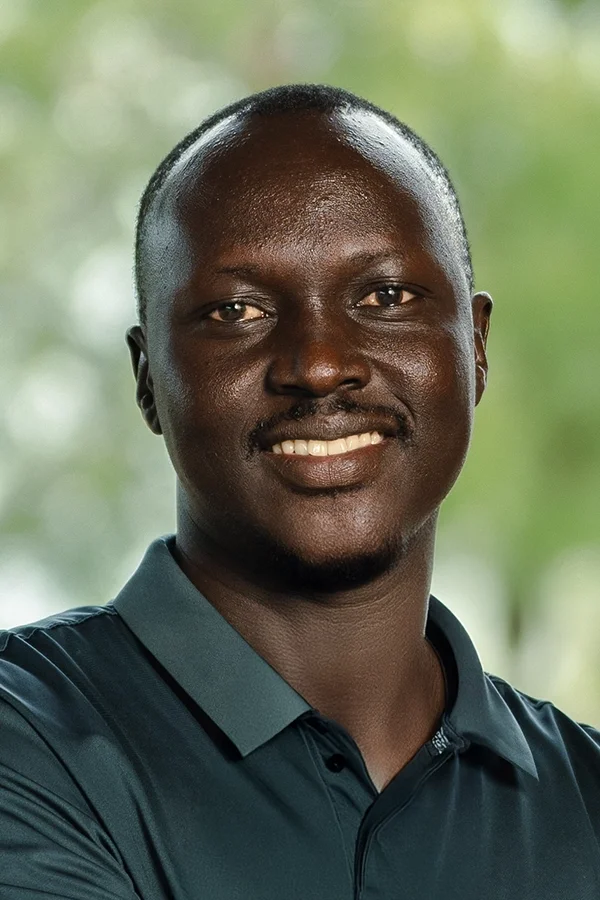 Portrait of a smiling man with short hair, wearing a dark blue collared shirt, outdoors with blurred green background.