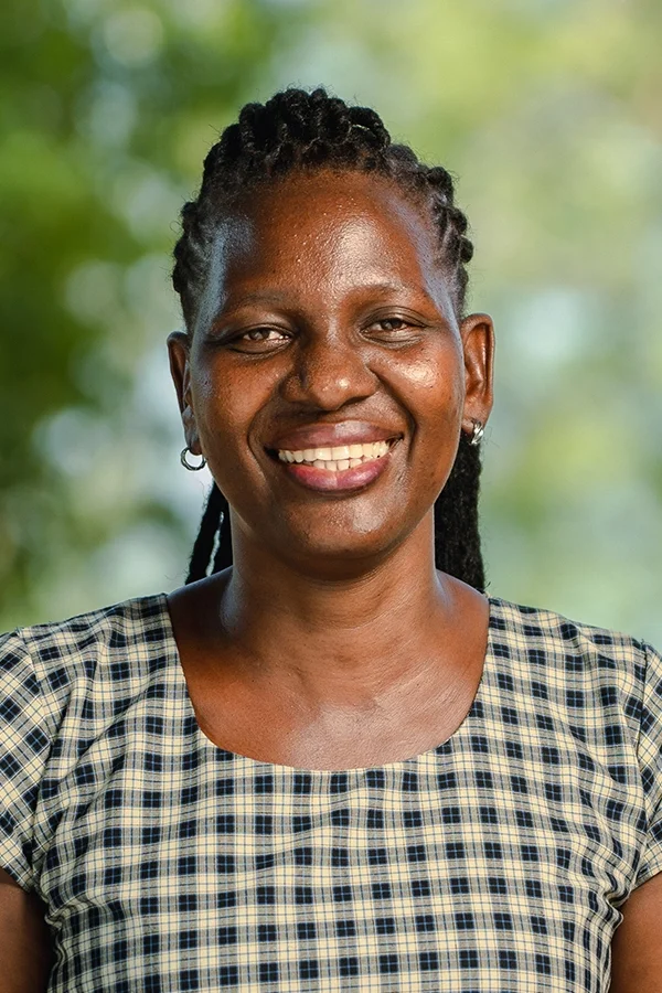 A woman smiling outdoors, wearing a checkered top, with a blurred green background.